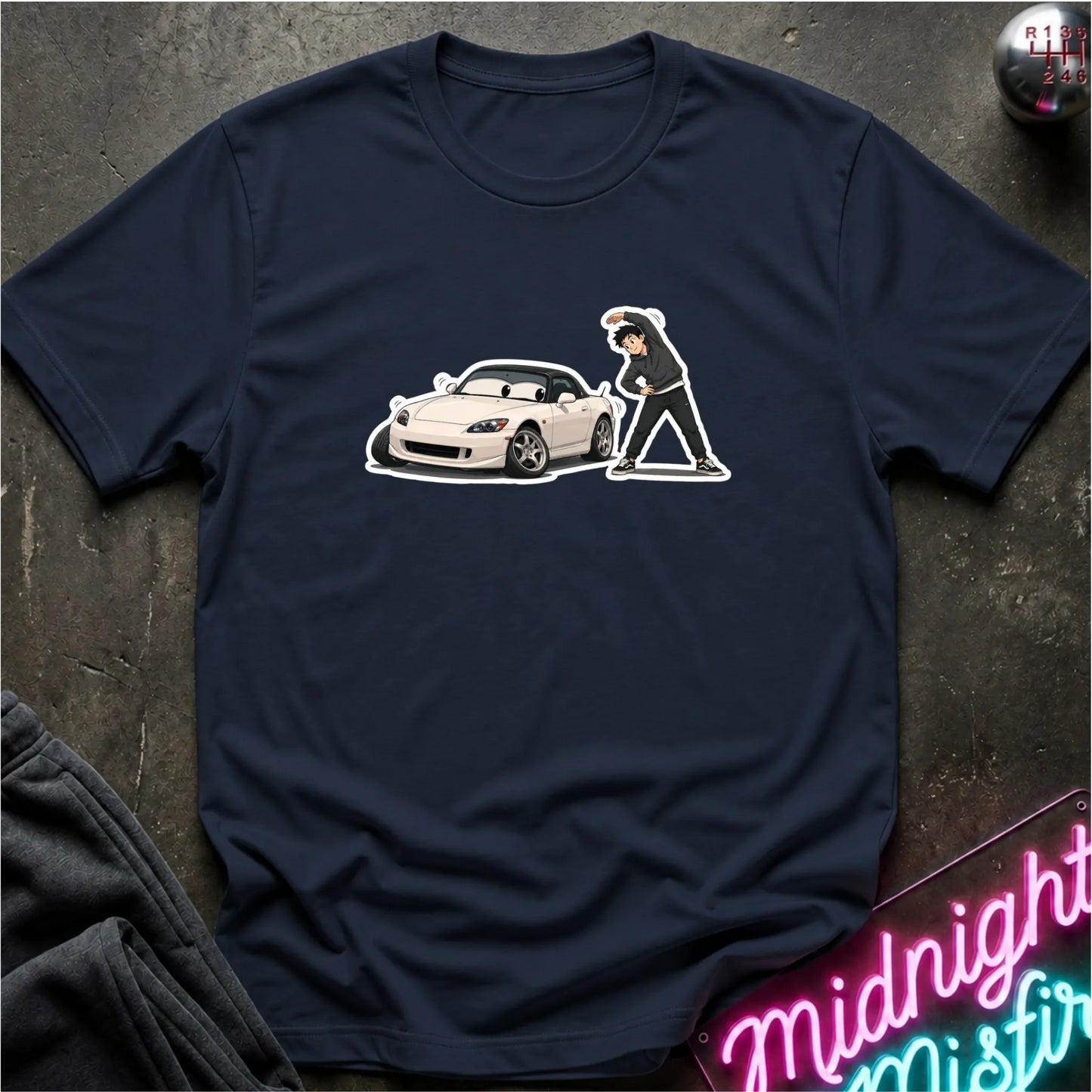 A navy blue crewneck t-shirt laid flat on a dark textured surface, featuring a graphic print of a white sports car and a person. The shirt is styled with a dark gray sweatshirt in the corner and a neon sign in the bottom right.