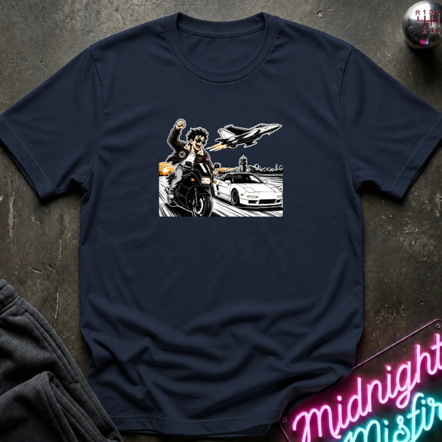 A navy short-sleeve crewneck t-shirt with a graphic print of a motorcycle racer, a sports car, and a jet plane, laid flat on a dark textured background with a neon sign and sweatpants partially visible.