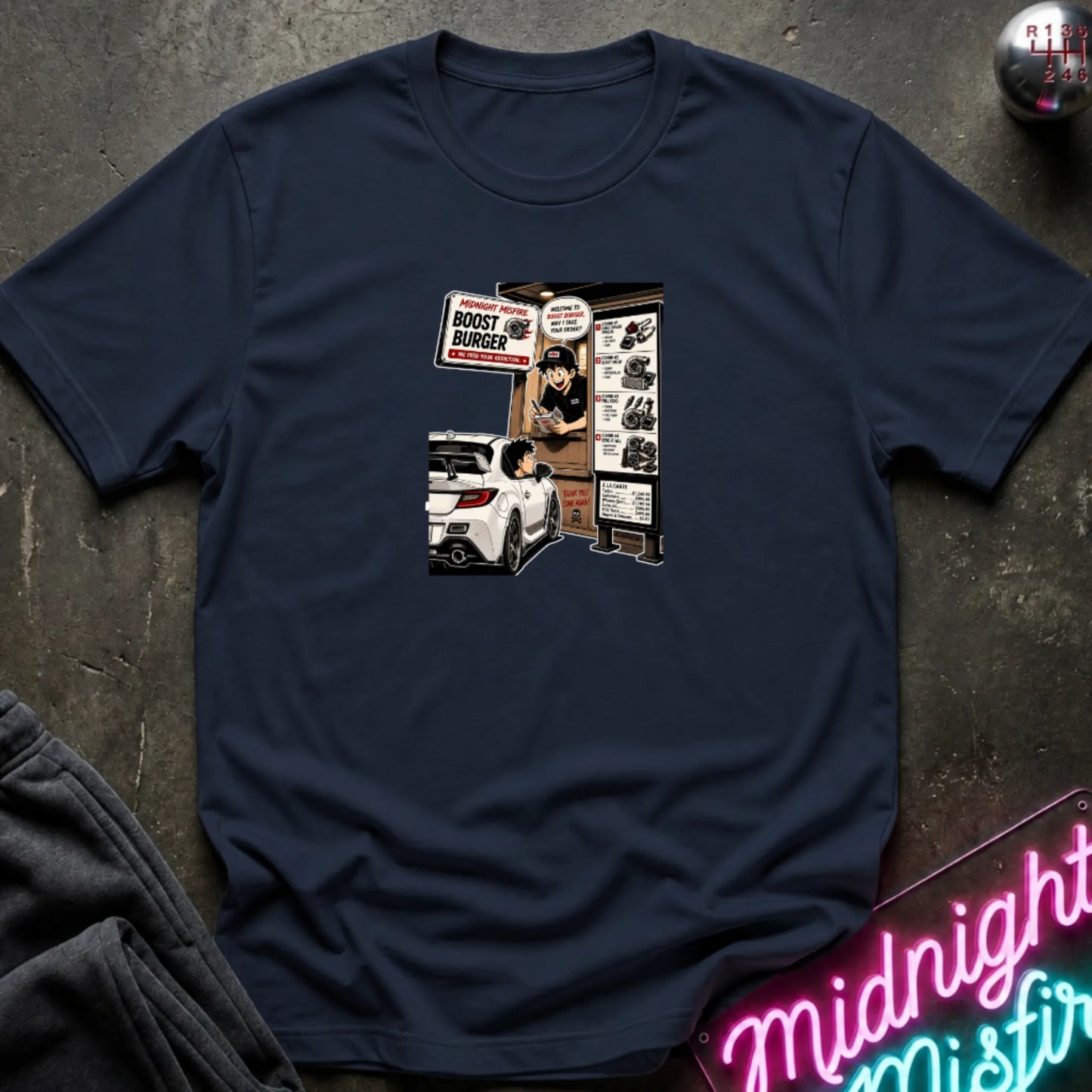 A navy  short-sleeve crewneck t-shirt laid flat on a dark textured surface, featuring a central graphic print of a street-food burger scene with a white car and signs. 