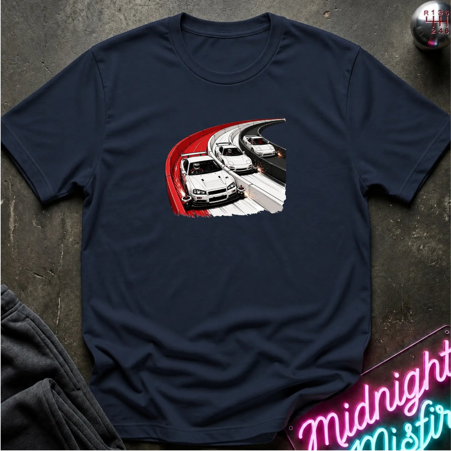 A navy blue short-sleeve t-shirt with a graphic print of three white sports cars racing on a highway, styled flat on a dark textured surface with a neon sign and pants partially visible.