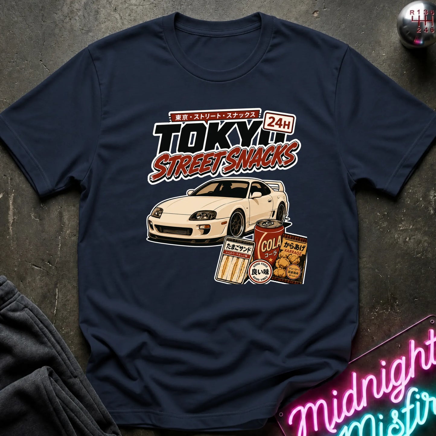 navy graphic t-shirt laid flat on a dark textured surface. The graphic features a white sports car and retro Japanese-style snack cans with the text 'TOKYO STREET SNACKS 24H'.