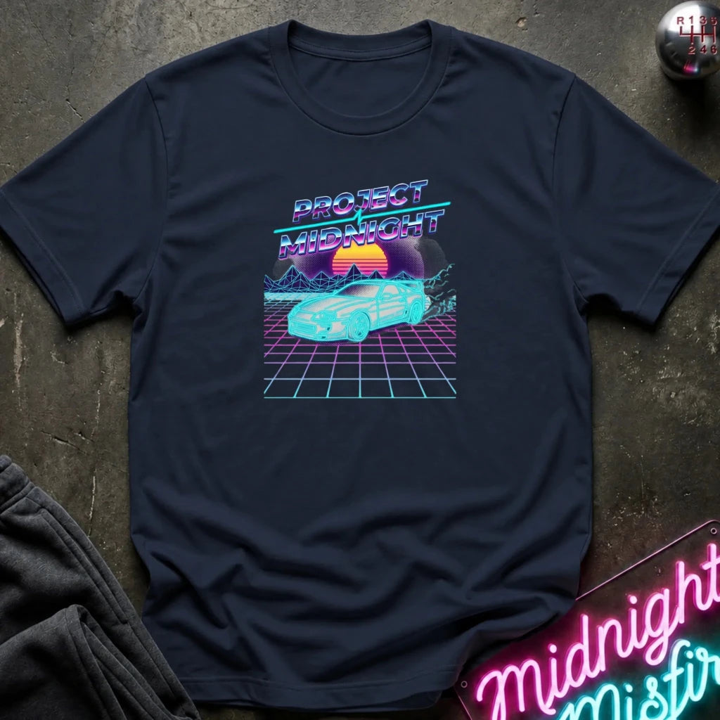 A navy blue short-sleeve t-shirt with a retro 80s synthwave graphic reading 'PROJECT MIDNIGHT' above a cyan car on a grid with a sunset, laid flat on a dark textured surface with a neon sign and folded fabric visible at the edges.