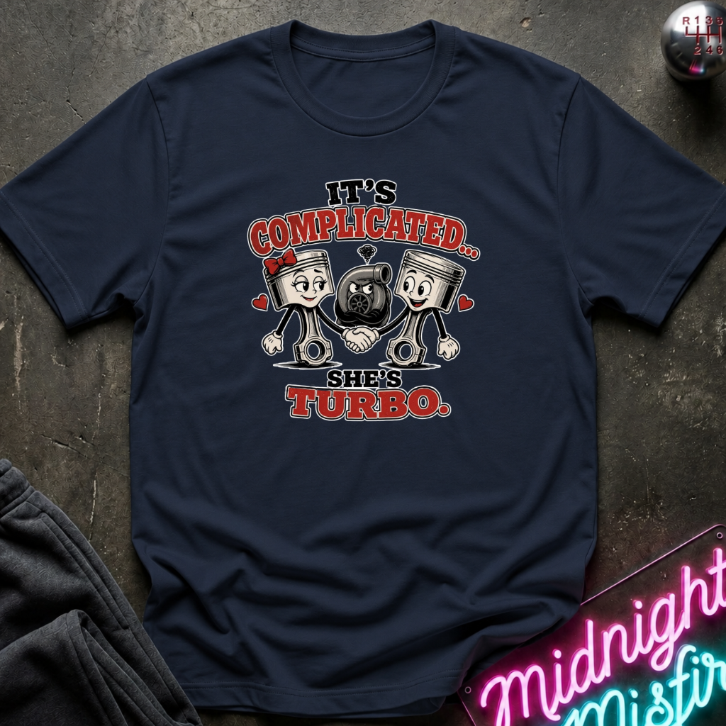 A navy blue graphic t-shirt laid flat on a dark textured surface. The shirt features a cartoon graphic of two pistons and a black turbo, with the text 'IT'S COMPLICATED.