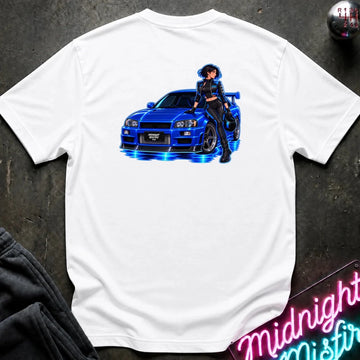 A white short-sleeve t-shirt laid flat on a dark textured surface, featuring a graphic print of a blue sports car with glowing blue lights and an anime-style woman standing beside it. Partially visible props include dark clothing in the lower left and a neon-style sign in the lower right.