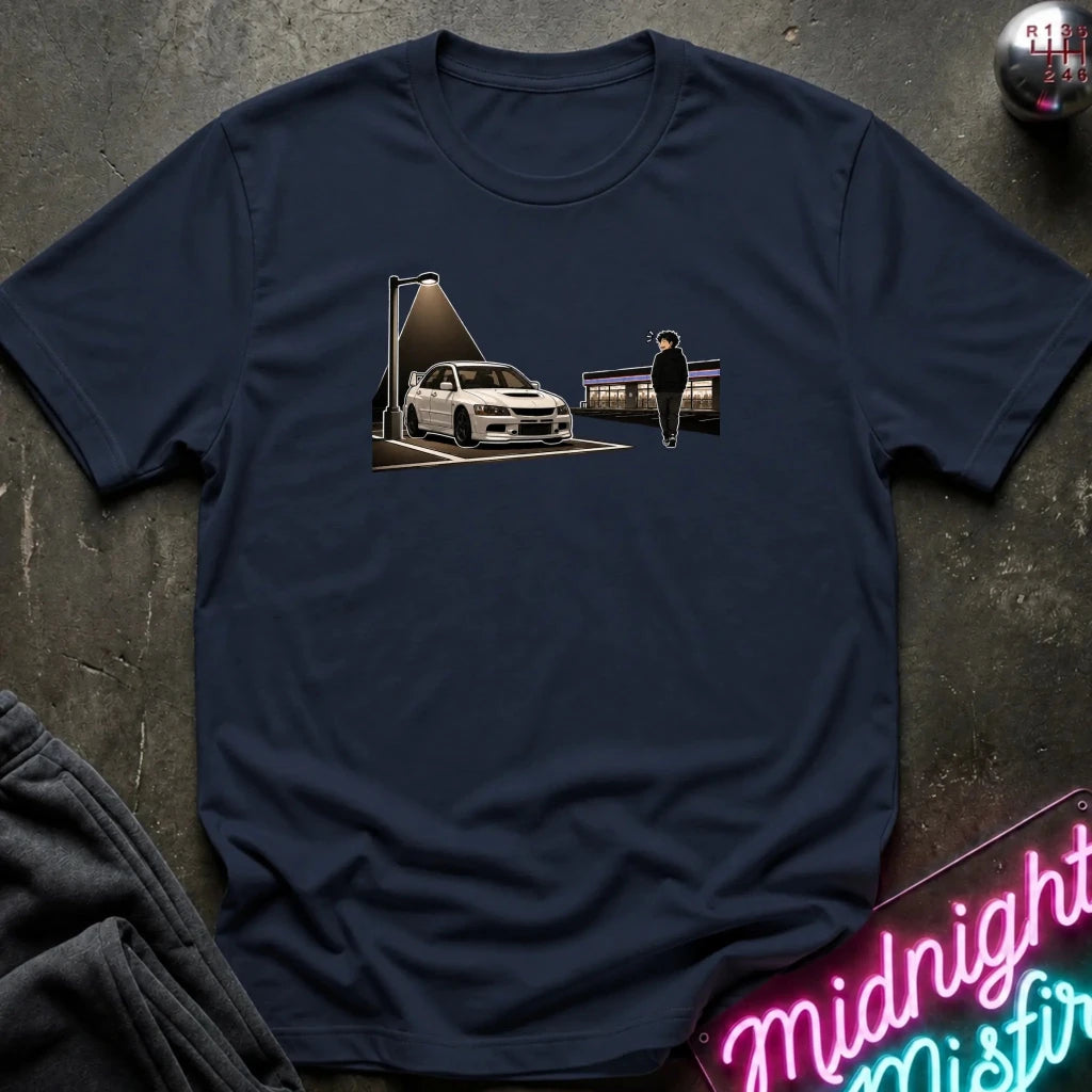 A navy blue short-sleeve t-shirt laid flat on a dark textured surface, featuring a graphic print of a white sports car under a street lamp with a silhouette figure standing nearby. 
