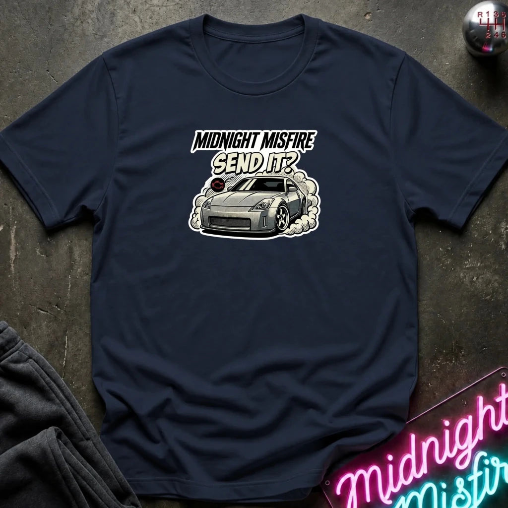 A navy blue graphic t-shirt laid flat on a dark textured surface. The shirt features a white and gray graphic of a sports car with smoke and the text 'MIDNIGHT MISFIRE SEND IT?'.