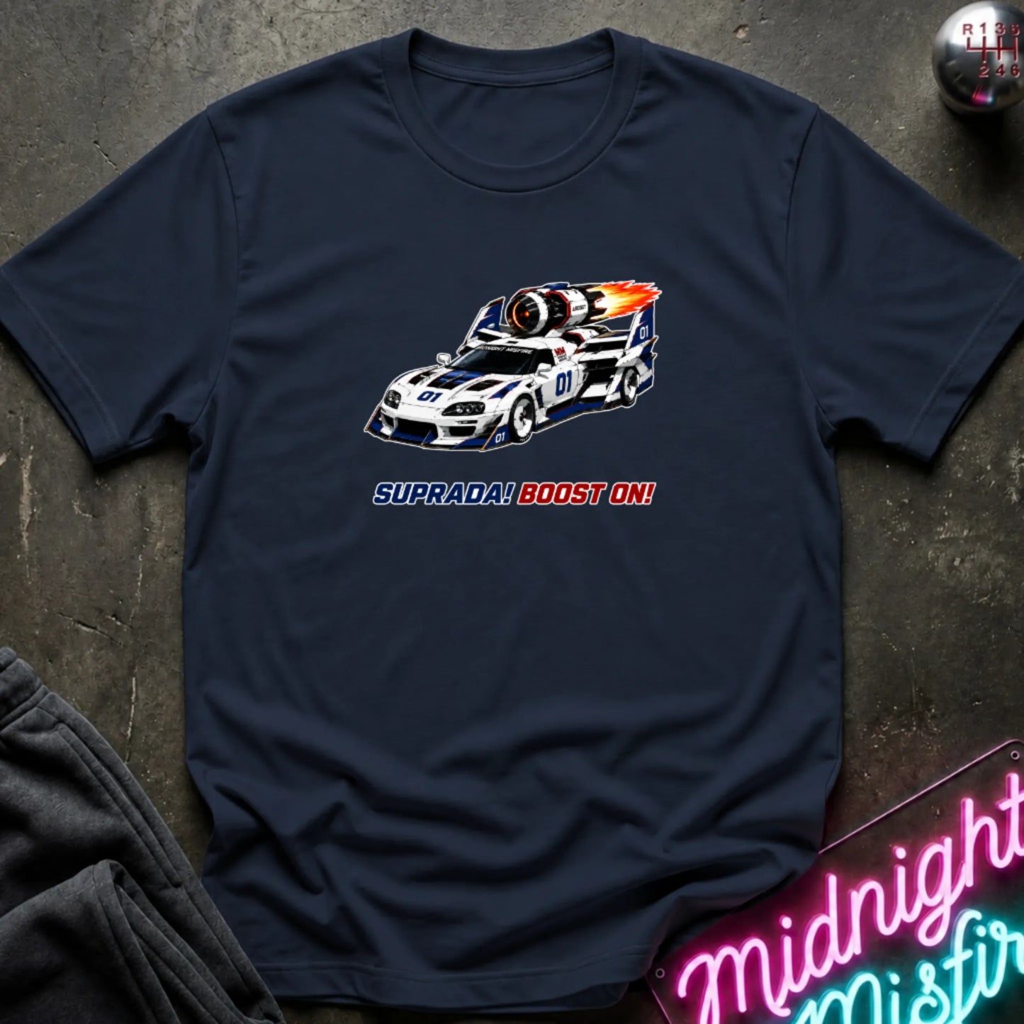 A navy short-sleeve t-shirt with a retro-style graphic of a race machine with big boost, displayed flat on a dark textured surface with a neon sign and sweatpants partially visible.