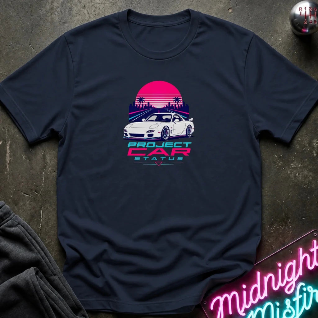 A navy blue short-sleeve t-shirt laid flat on a dark textured surface, featuring a retro-style graphic of a white sports car in front of a pink and purple sunset with the text 'PROJECT CAR STATUS'.