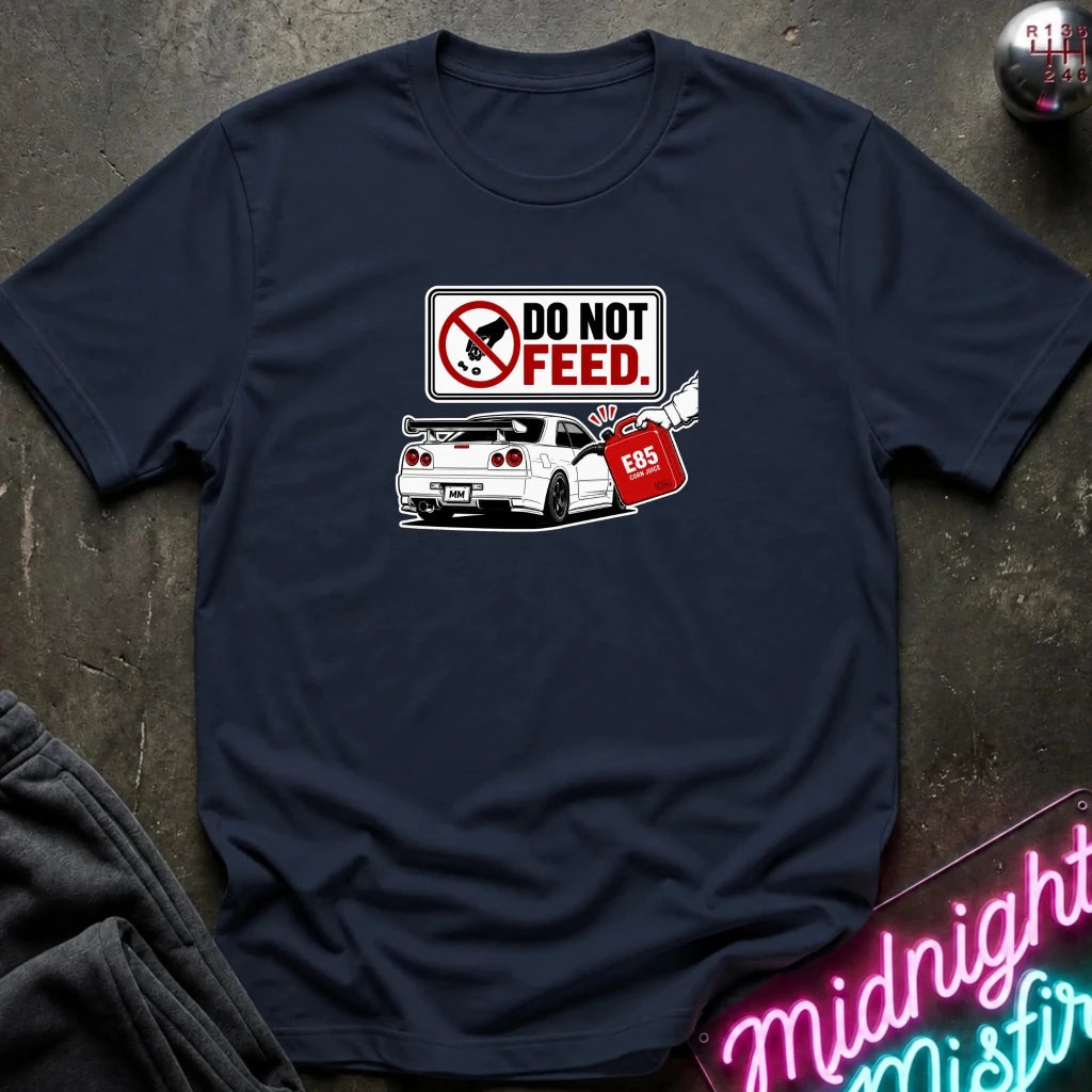 A navy short-sleeve graphic t-shirt laid flat on a dark textured surface. The graphic features a 'DO NOT FEED' sign.