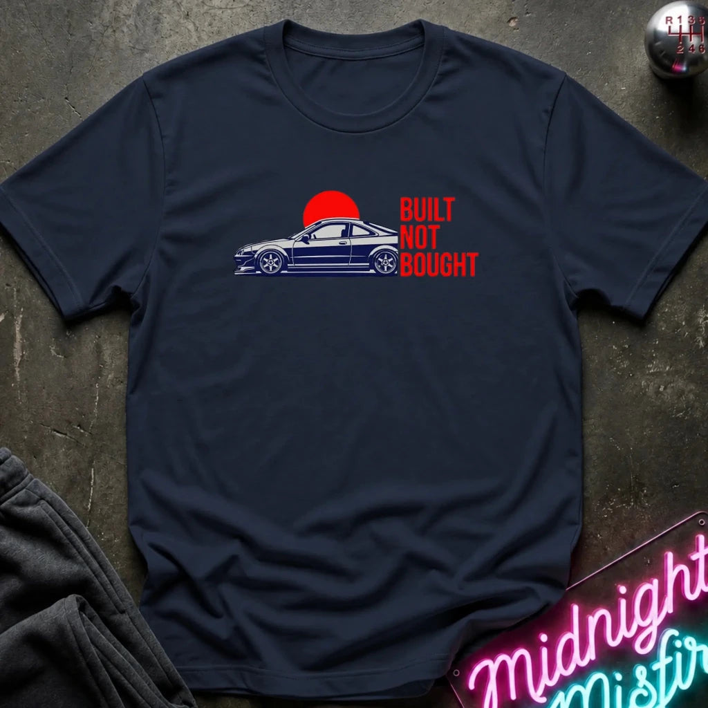 A navy blue graphic t-shirt laid flat on a dark textured surface. The shirt features a graphic of a blue sports car in front of a red sun with the text 'BUILT NOT BOUGHT' in red.
