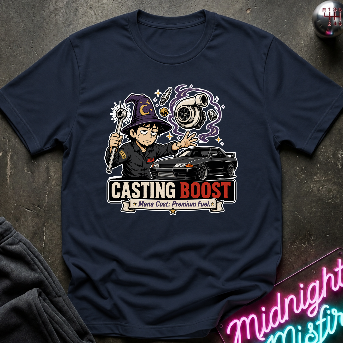 A navy blue graphic t-shirt laid flat on a dark textured surface. The graphic features a cartoon wizard holding a wand next to a black sports car, with the text 'CASTING BOOST' and 'Mana Cost: Premium Fuel'.
