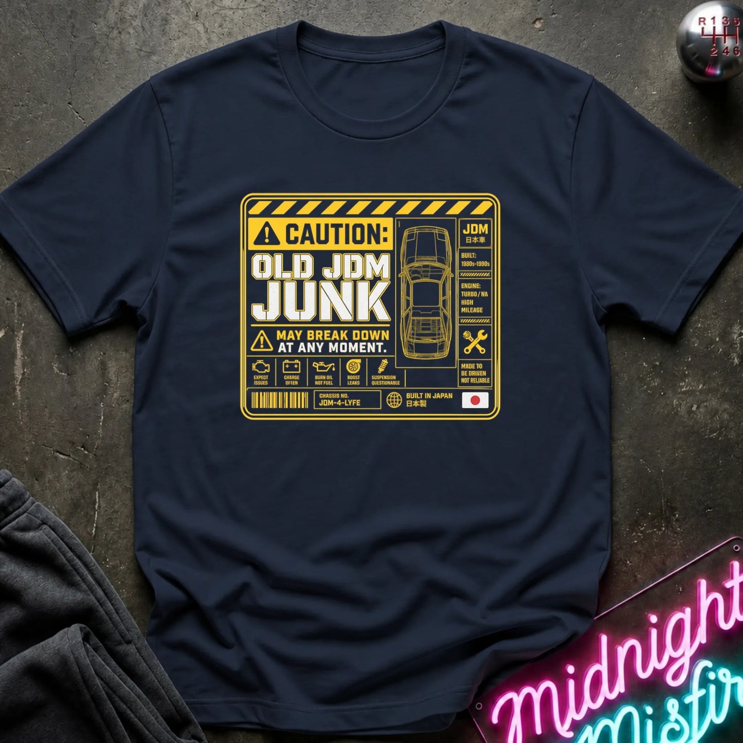 A navy blue short-sleeve t-shirt with a yellow and white graphic print on the front. The graphic features a caution-style warning label with the text 'CAUTION: OLD JDM JUNK MAY BREAK DOWN AT ANY MOMENT.
