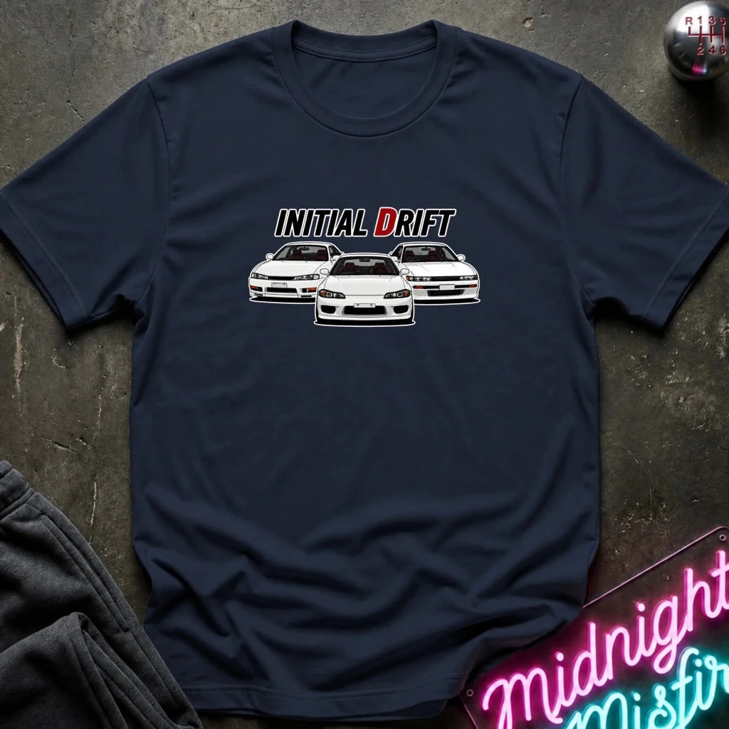 A navy blue short-sleeve t-shirt laid flat on a dark textured surface, featuring a graphic of three white drift cars with the text 'INITIAL DRIFT' above them. A neon sign and folded dark fabric are partially visible in the corners as props.