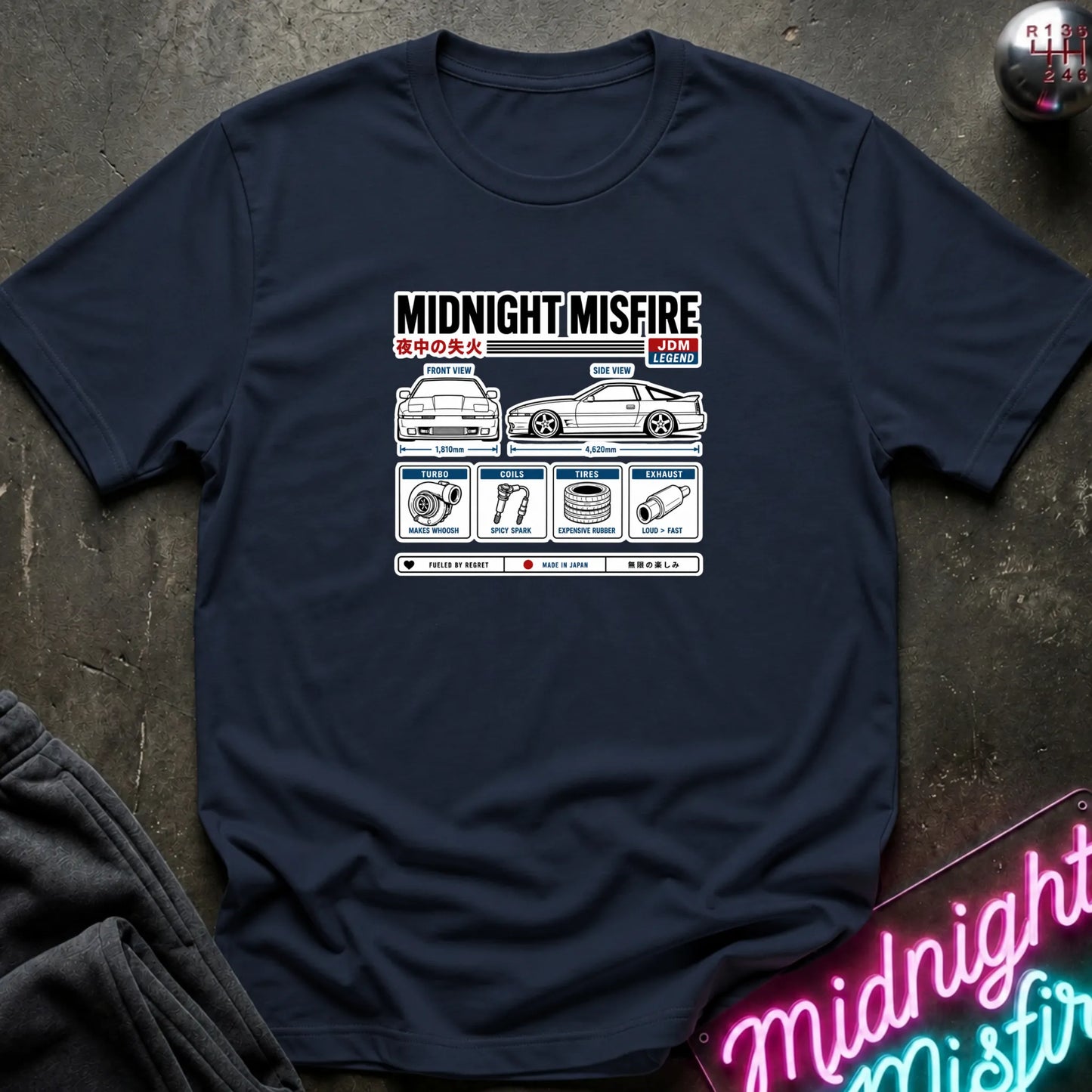 A navy blue short-sleeve t-shirt with a white and red graphic print on the chest, laid flat on a dark textured surface with a neon sign and dark fabric in the corners.