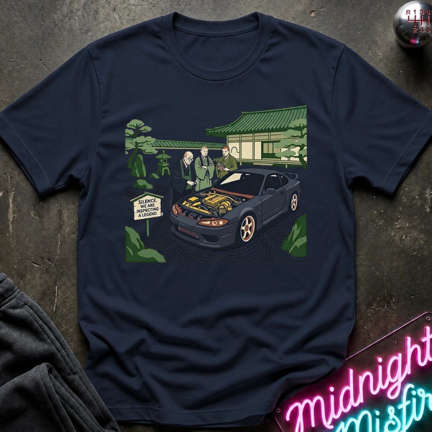 Navy short-sleeve graphic t-shirt laid flat on a dark textured surface. The graphic shows a black sports car with its hood open, surrounded by two men in suits, a Japanese-style building, and green trees.