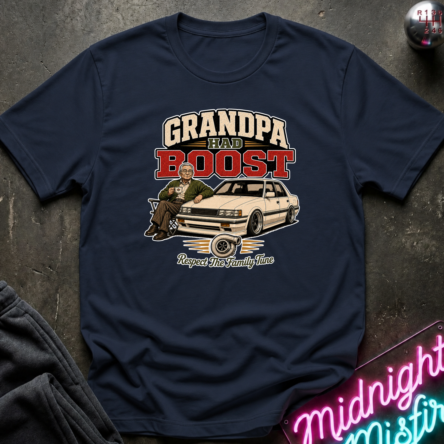 A navy blue graphic t-shirt laid flat on a dark textured surface. The shirt features a vintage-style graphic with the text 'GRANDPA HAD BOOST' in cream and red, an illustration of a man sitting on a white classic car, and the phrase 'Respect The Family Line' below.