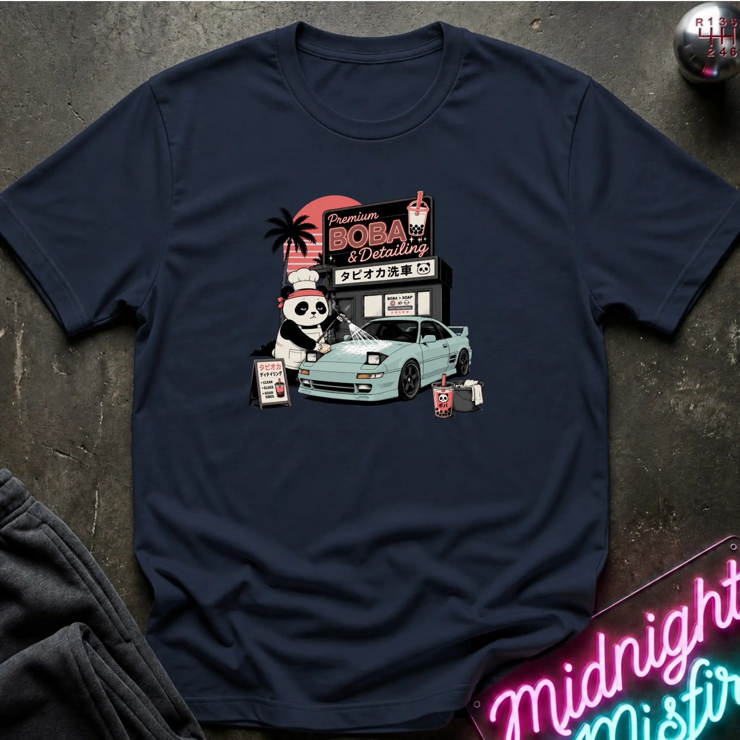 A navy blue short-sleeve t-shirt with a colorful graphic print on the chest, laid flat on a dark textured surface with a neon sign and dark pants partially visible in the corners.