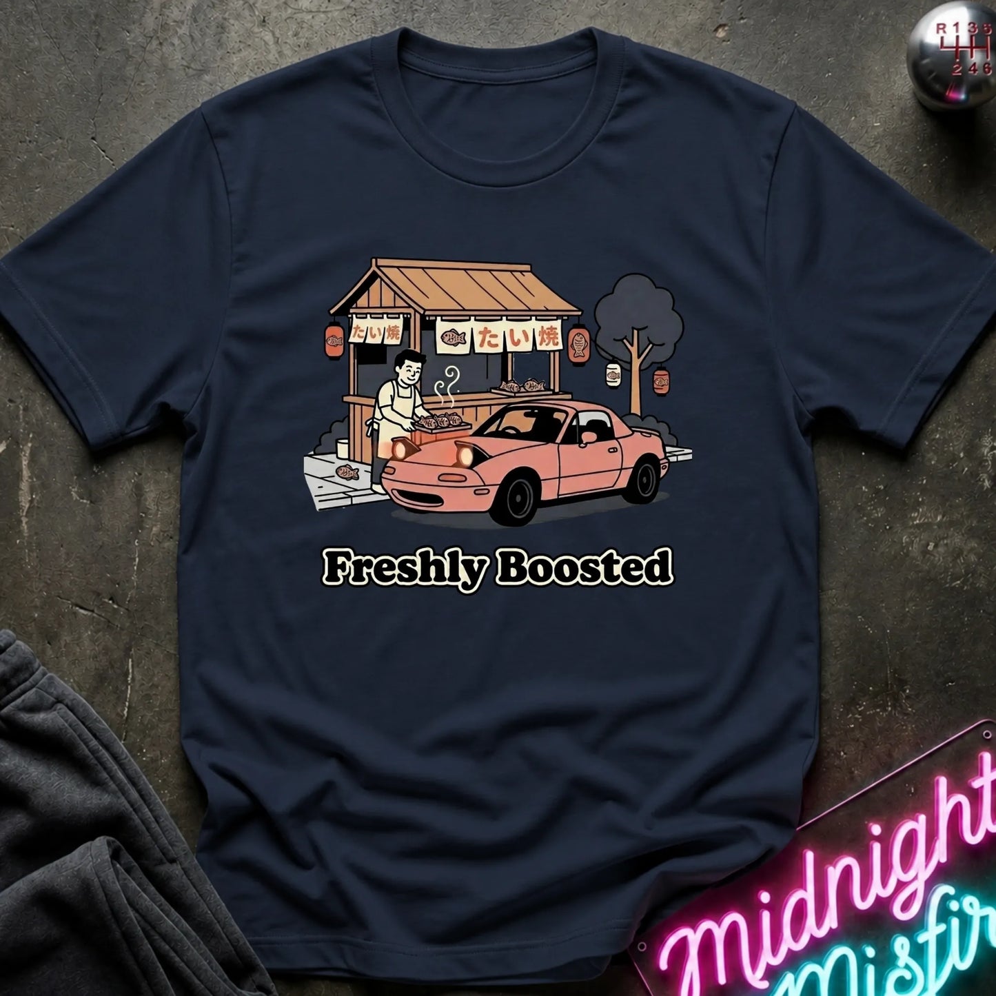 A navy blue graphic t-shirt laid flat on a dark textured surface. The graphic features a pink convertible car parked in front of a street food stall  and the text 'Freshly Boosted' printed below.