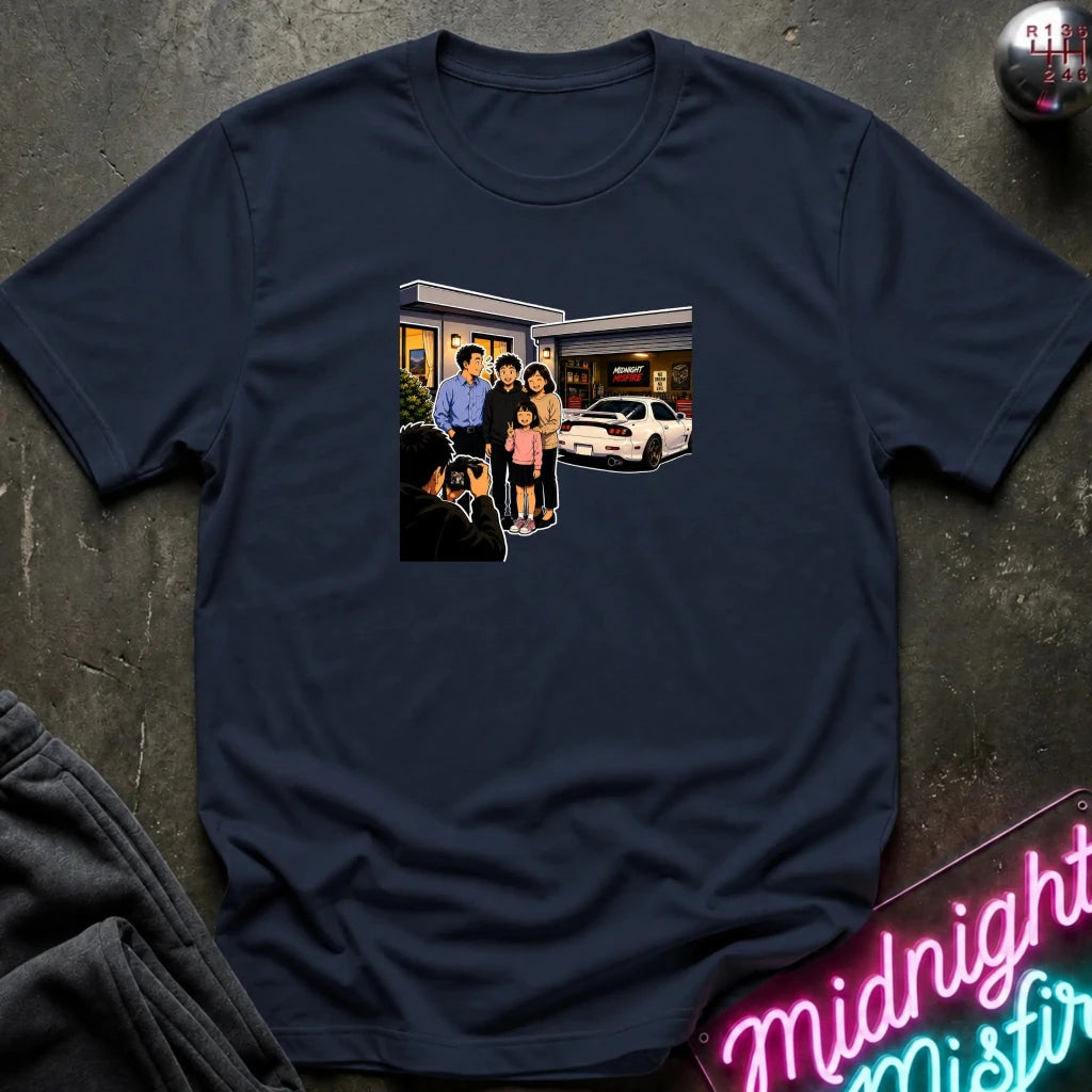 A navy blue short-sleeve crewneck t-shirt laid flat on a dark textured surface, featuring a rectangular graphic print of a family standing outside a building taking a photo.