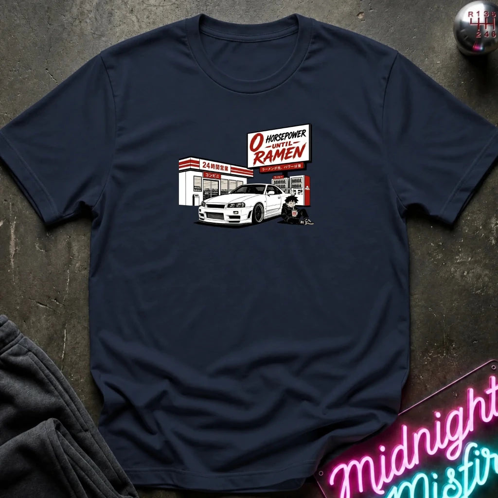A navy blue crewneck t-shirt laid flat on a dark textured surface, featuring a graphic print of a white sports car parked in front of a konbini.