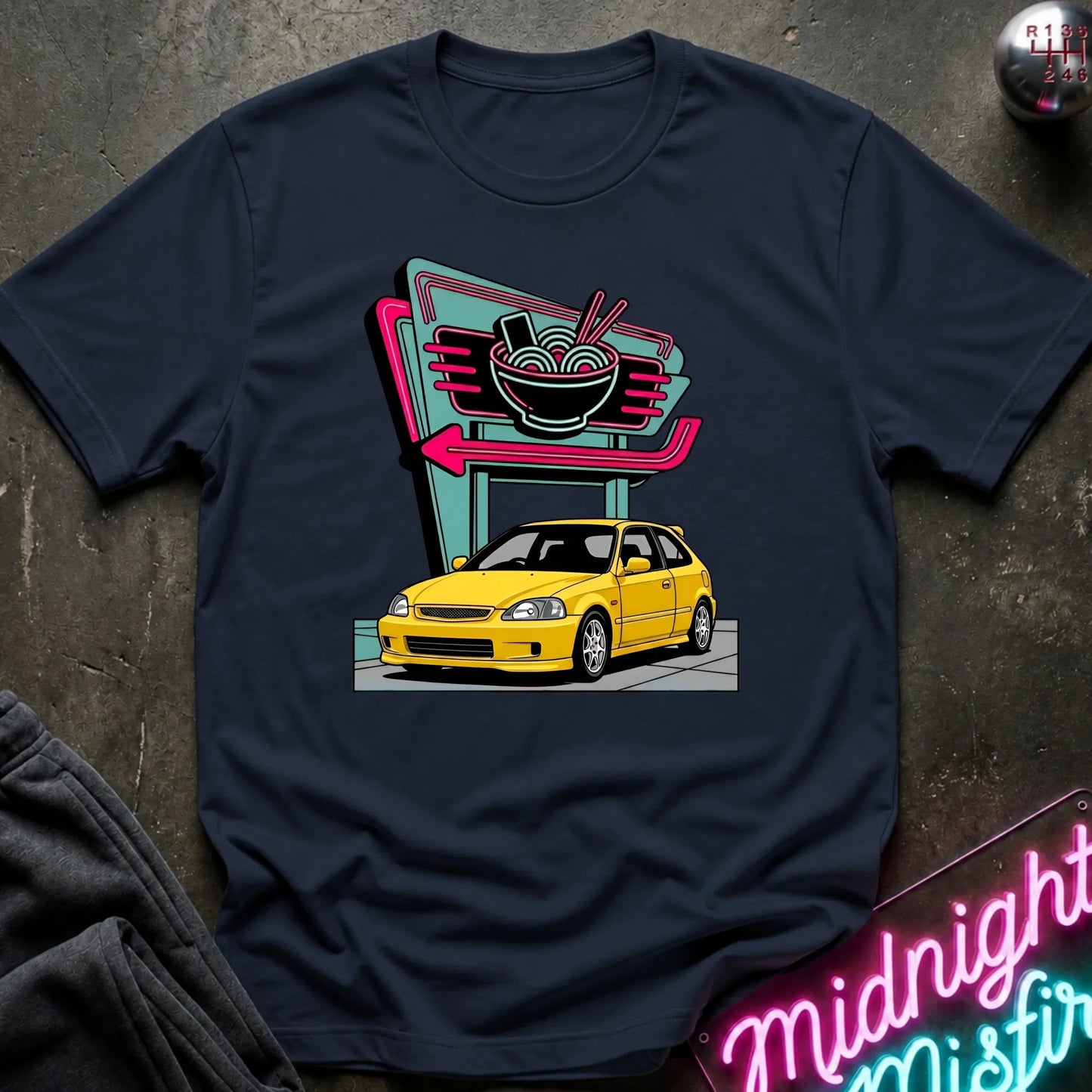 A navy blue graphic t-shirt laid flat on a dark textured surface. The graphic features a retro neon sign with a bowl of ramen and an arrow pointing to a yellow car.