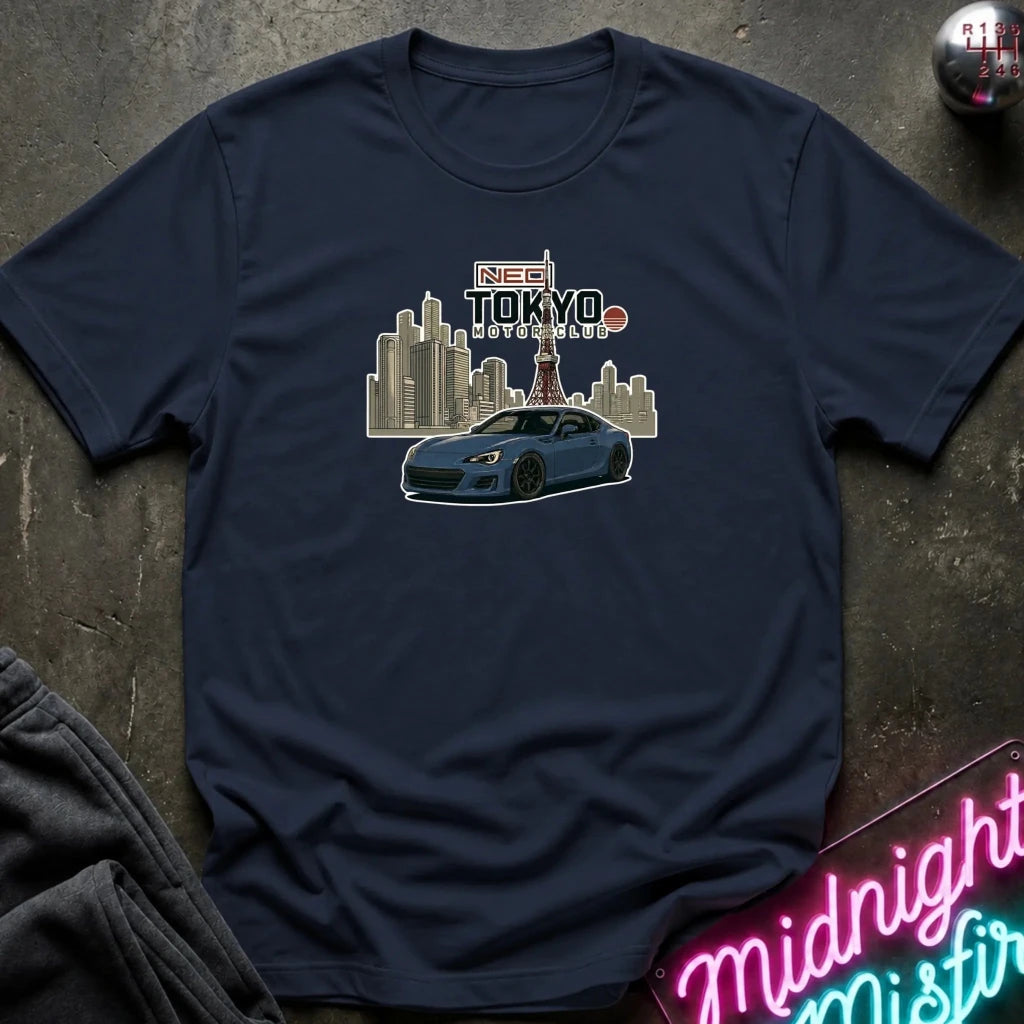 A navy blue short-sleeve t-shirt laid flat on a dark textured surface, featuring a graphic print of a blue sports car in front of a Tokyo skyline with the text 