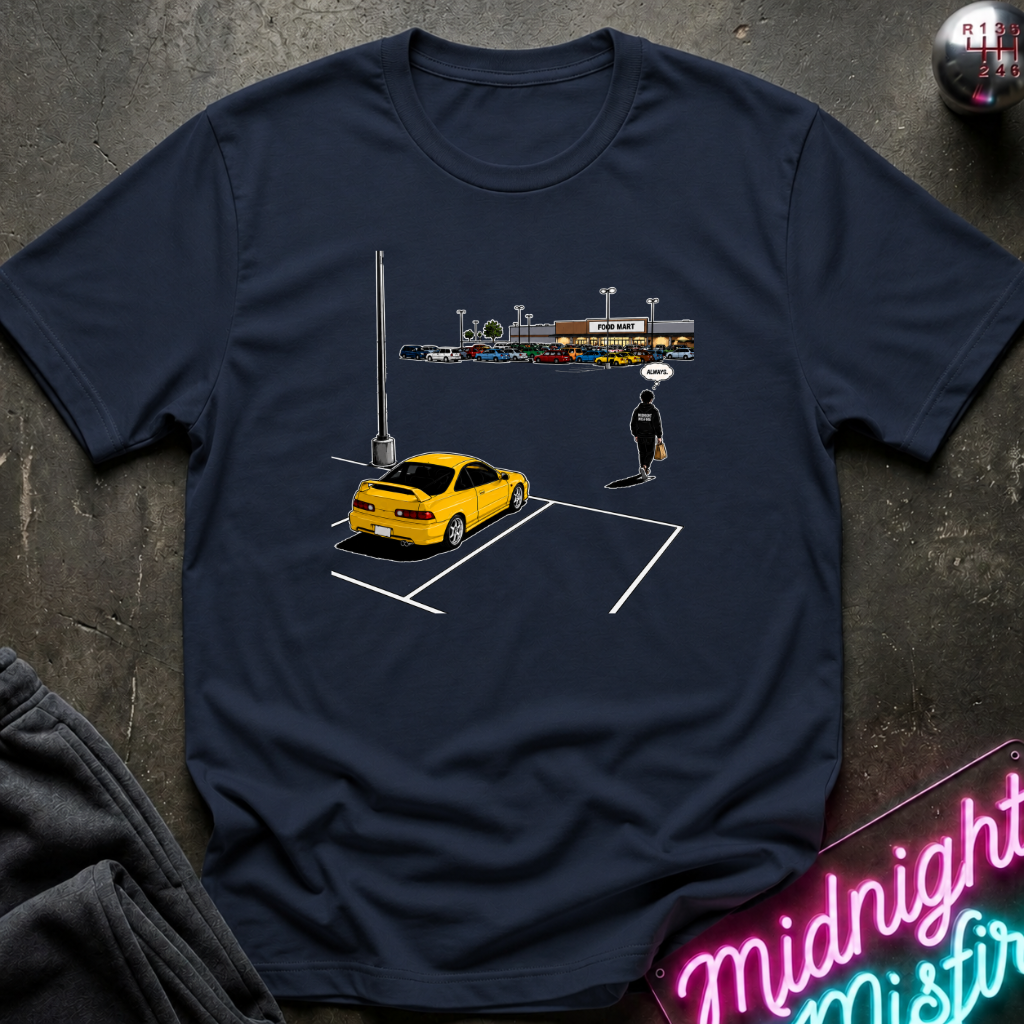 A navy blue short-sleeve t-shirt with a graphic print of a yellow sports car in a parking spot and a small figure, laid flat on a dark textured surface with a neon sign and sweatpants partially visible.