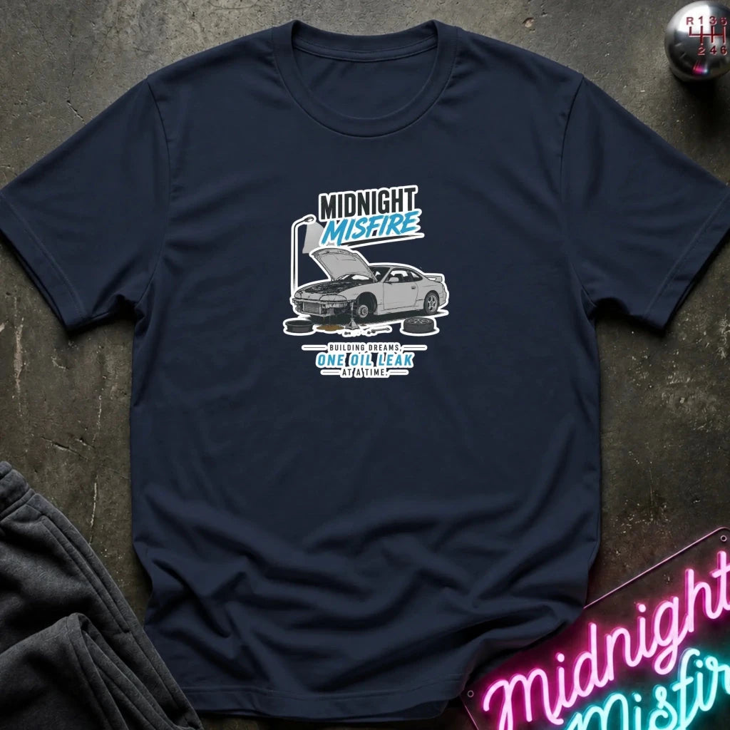A navy blue short-sleeve t-shirt laid flat on a dark textured surface, featuring a graphic print of a silver sports car with its hood open and the text 'Midnight Misfire' 