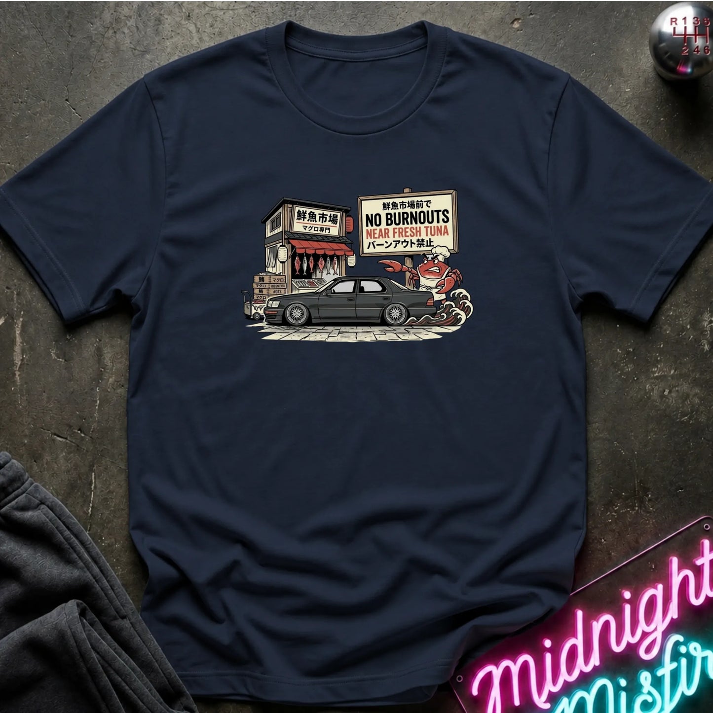A navy blue short-sleeve t-shirt with a vintage-style graphic print on the chest, laid flat on a dark textured surface with decorative props.