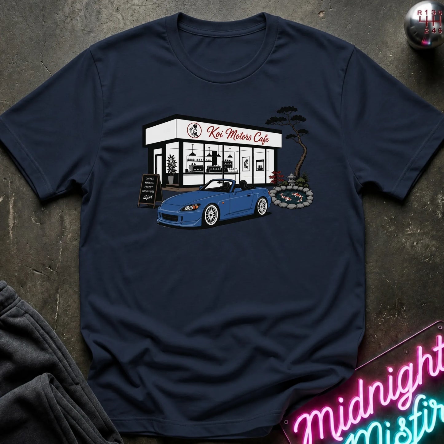 A navy blue short-sleeve t-shirt laid flat on a dark textured surface, featuring a graphic print of a blue convertible sports car parked in front of a cafe named 'Koi Motor Cafe'.