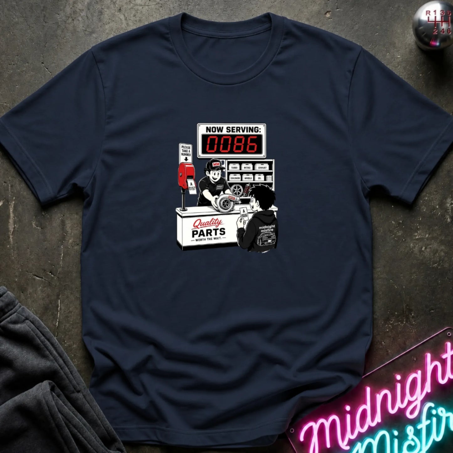 A navy blue short-sleeve t-shirt with a retro-style graphic of a garage attendant at a 'Quality Parts' counter, displayed flat on a dark textured surface with a neon sign and sweatpants partially visible.