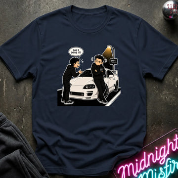 A navy blue graphic t-shirt laid flat on a dark textured surface. The shirt features a cartoon illustration of a man in a black hoodie asking if he can drive the white sports car next to him, with a speech bubble reading 'CAN I DRIVE IT?'.