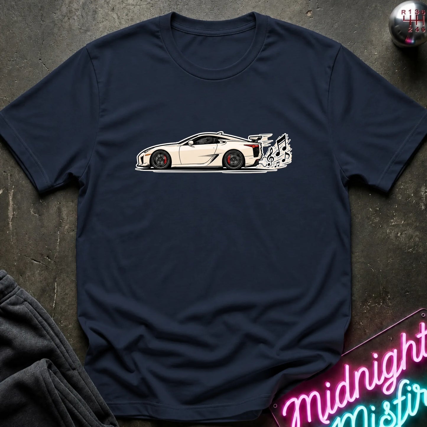A navy blue short-sleeve t-shirt laid flat on a dark textured surface, featuring a graphic print of a white sports car with a music note coming from exhaust.