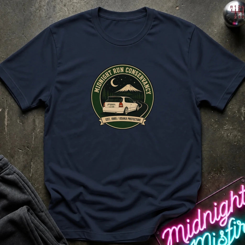 A navy blue short-sleeve t-shirt laid flat on a dark textured surface, featuring a circular vintage-style graphic on the chest with a white car, mountain, moon, and the words 'Midnight Run Conservancy'