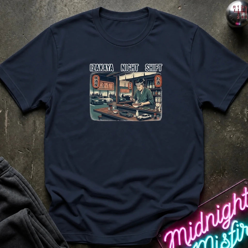A navy blue graphic t-shirt laid flat on a dark textured surface. The shirt features a rectangular vintage-style illustration of a man working late at a diner, with the text 'IZAKAYA NIGHT SHIFT' printed above it.