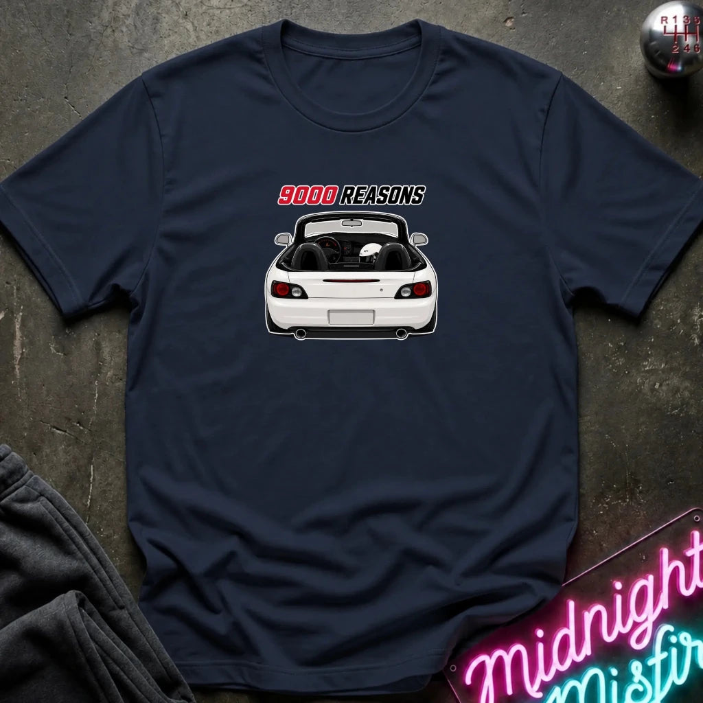A navy blue short-sleeve t-shirt laid flat on a dark textured surface. The shirt features a graphic print of a white convertible sports car with the text '9000 REASONS' above it in red and white.