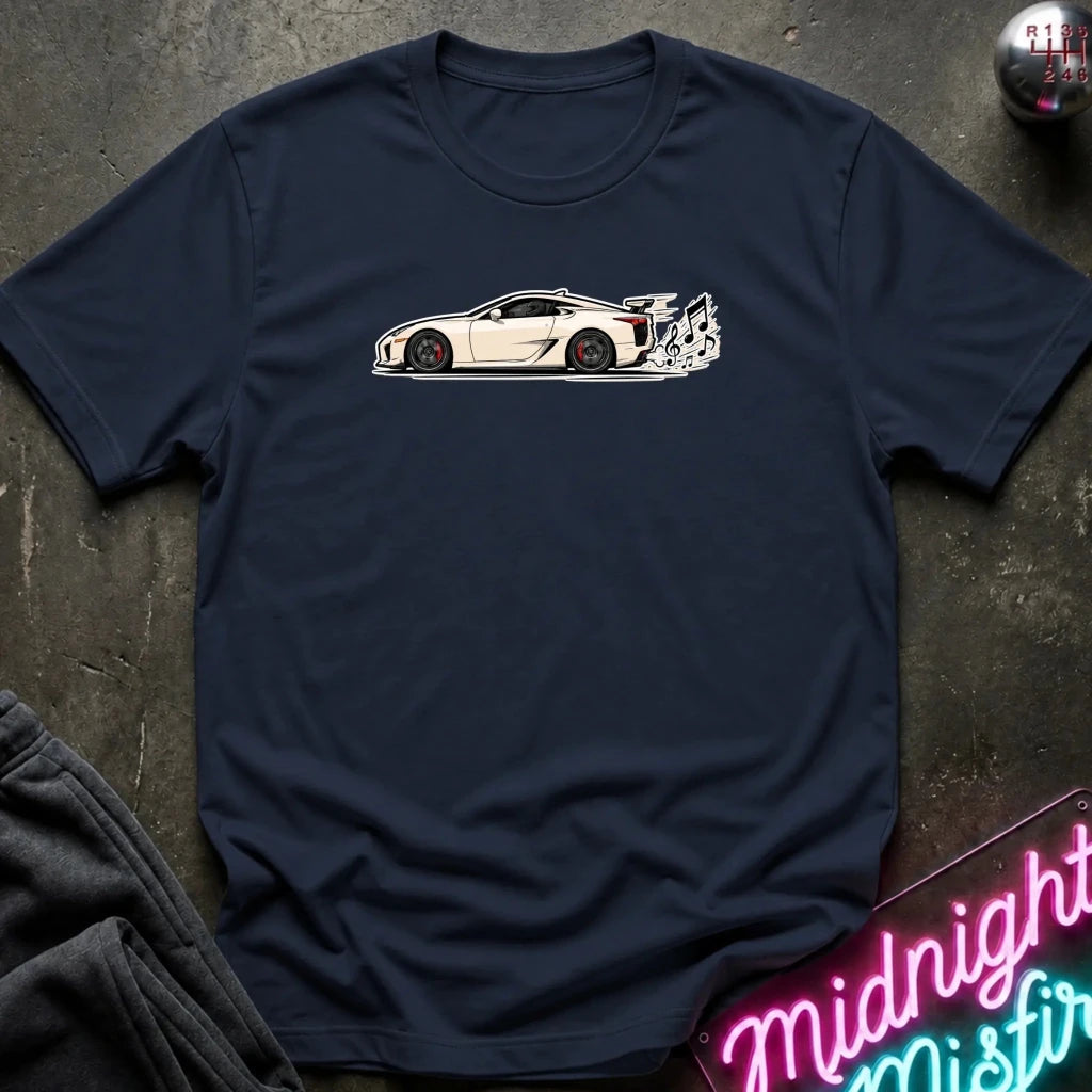 A navy blue short-sleeve t-shirt laid flat on a dark textured surface, featuring a white side-profile graphic of a sports car with a rear spoiler and exhaust flames. A folded dark sweatshirt corner and a pink neon sign are partially visible as props.