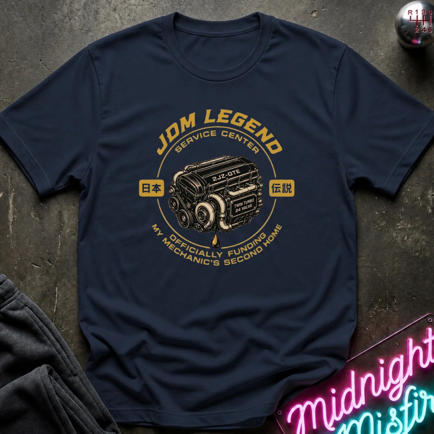 A navy blue graphic t-shirt laid flat on a dark textured surface. The shirt features a circular yellow and black graphic of a car engine with Japanese text and the words 'JDM LEGEND SERVICE CENTER' and 'MY MECHANIC'S SECOND HOME'.