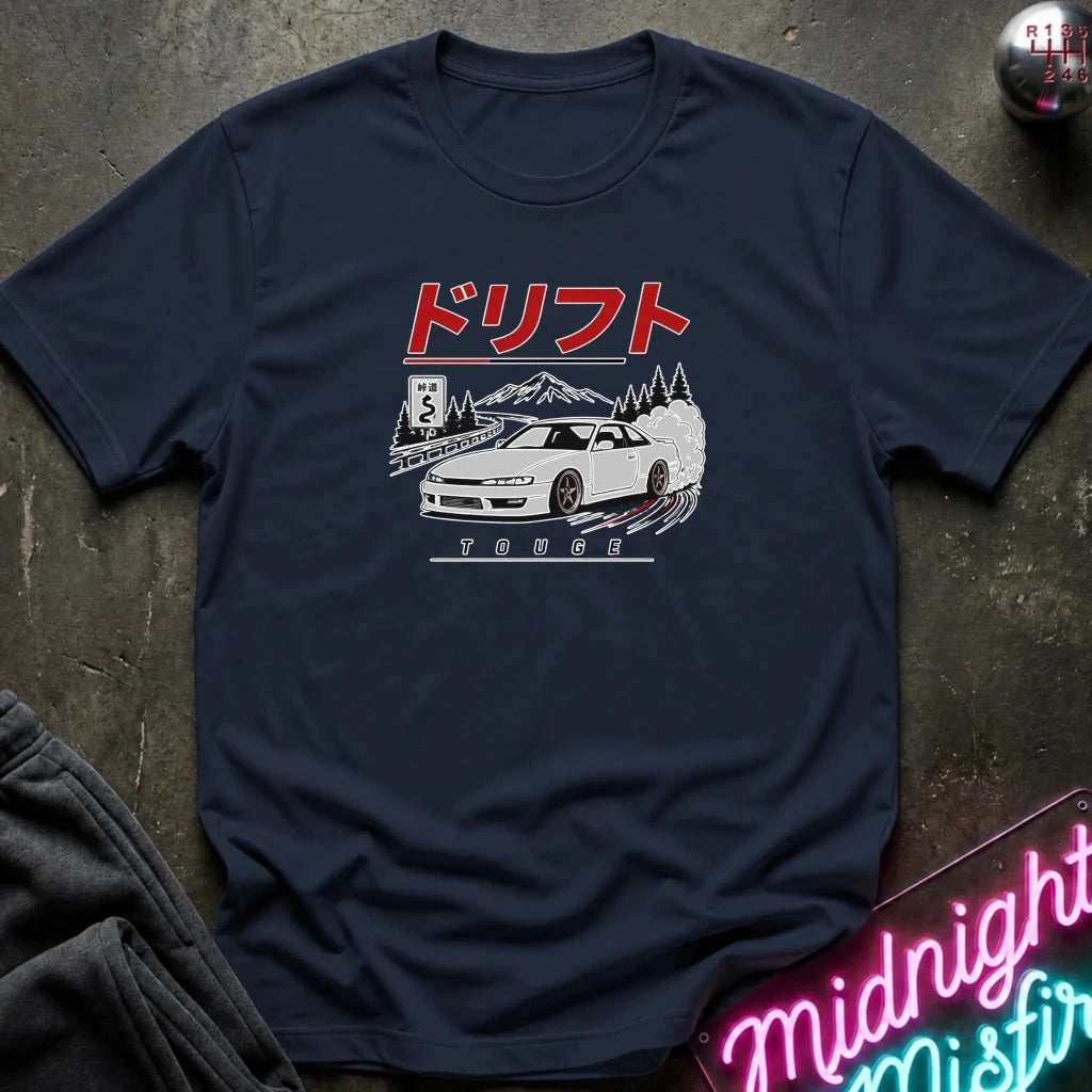 A navy blue short-sleeve t-shirt with a graphic print of a white sports car, mountains, and Japanese text, laid flat on a dark textured surface with a neon sign and dark pants partially visible.