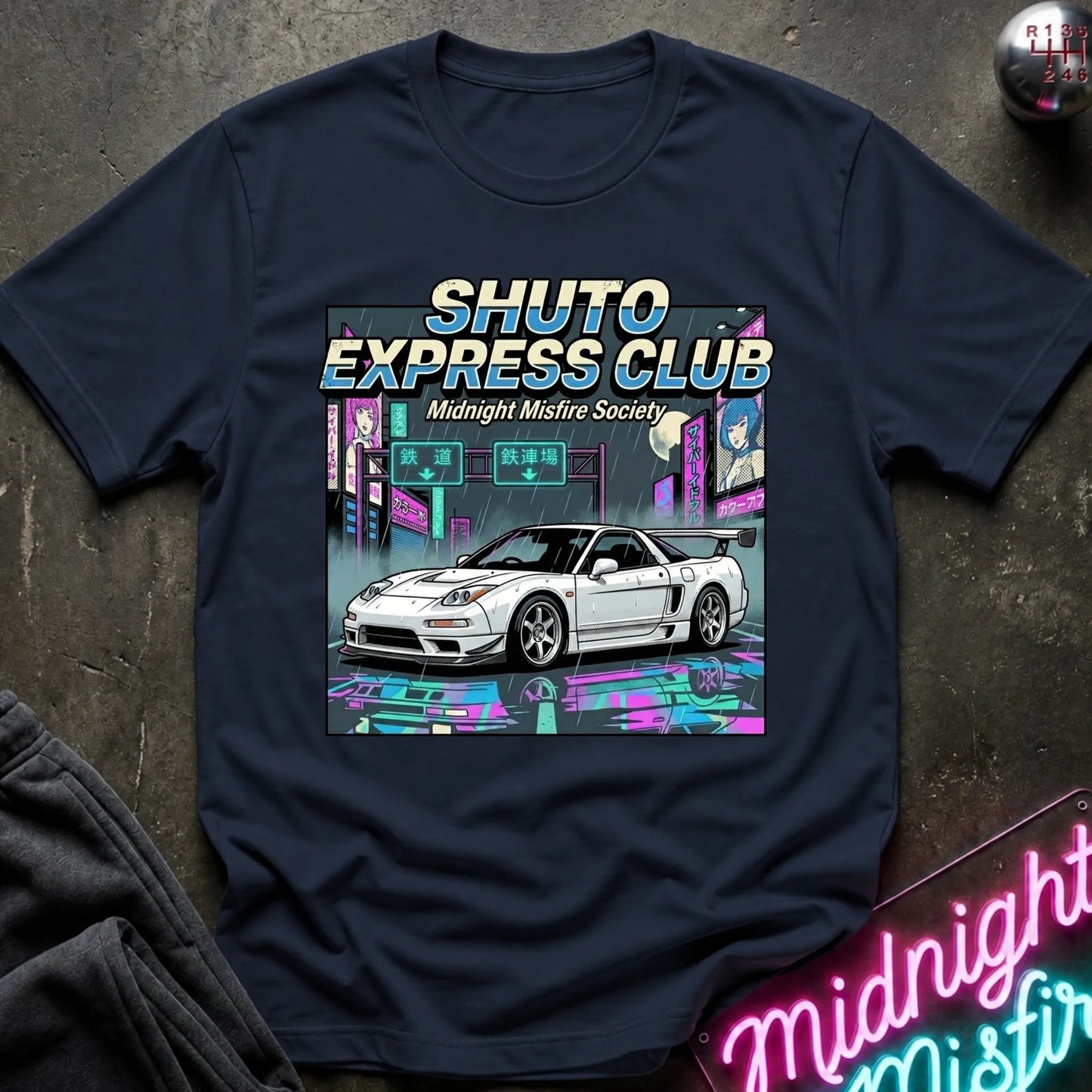 A navy blue graphic t-shirt laid flat on a dark textured surface. The shirt features a colorful graphic print of a white sports car in front of a neon cyberpunk city background, with the text 'SHUTO EXPRESS CLUB' and 'Midnight Misfire Society'.