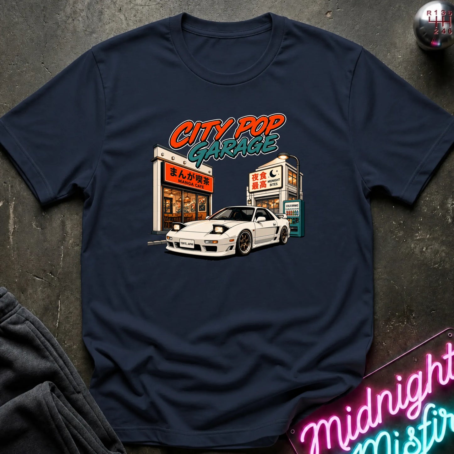 A navy blue short-sleeve graphic t-shirt laid flat on a dark textured surface. The graphic features a white sports car parked in front of a cafe with the text 'CITY POP GARAGE' in orange and teal.