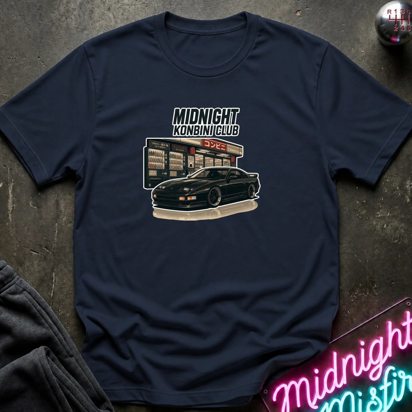 A navy blue short-sleeve t-shirt with a vintage-style graphic of a black sports car in front of a convenience store reading 'Midnight Kombi Club' and 'Open 24 Hours'. The shirt is laid flat on a dark textured surface with a neon sign and folded dark fabric partially visible.