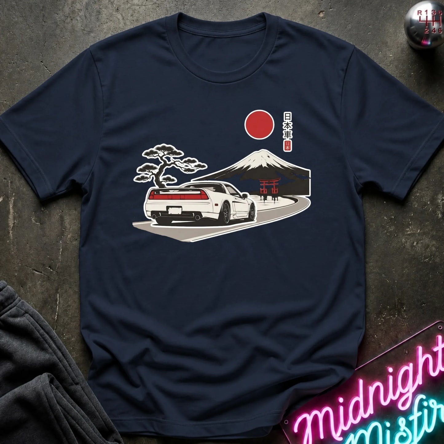A navy blue short-sleeve t-shirt laid flat on a dark textured surface, featuring a graphic print of a white sports car on a road with a red sun, Mount Fuji, a black pine tree, and a red torii gate, alongside Japanese text.
