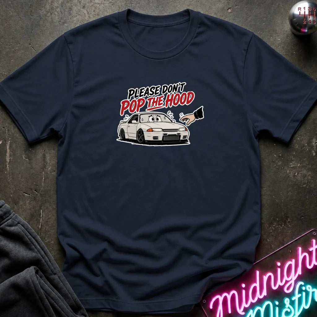 A navy blue graphic t-shirt laid flat on a dark textured surface. The shirt features a white car illustration and the text 'PLEASE DON'T POP THE HOOD' in white and red.