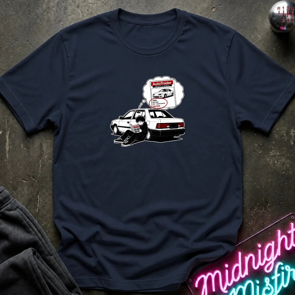 A navy blue short-sleeve t-shirt laid flat on a dark textured surface, featuring a graphic of a black-and-whitecar with a thought bubble showing a car part 