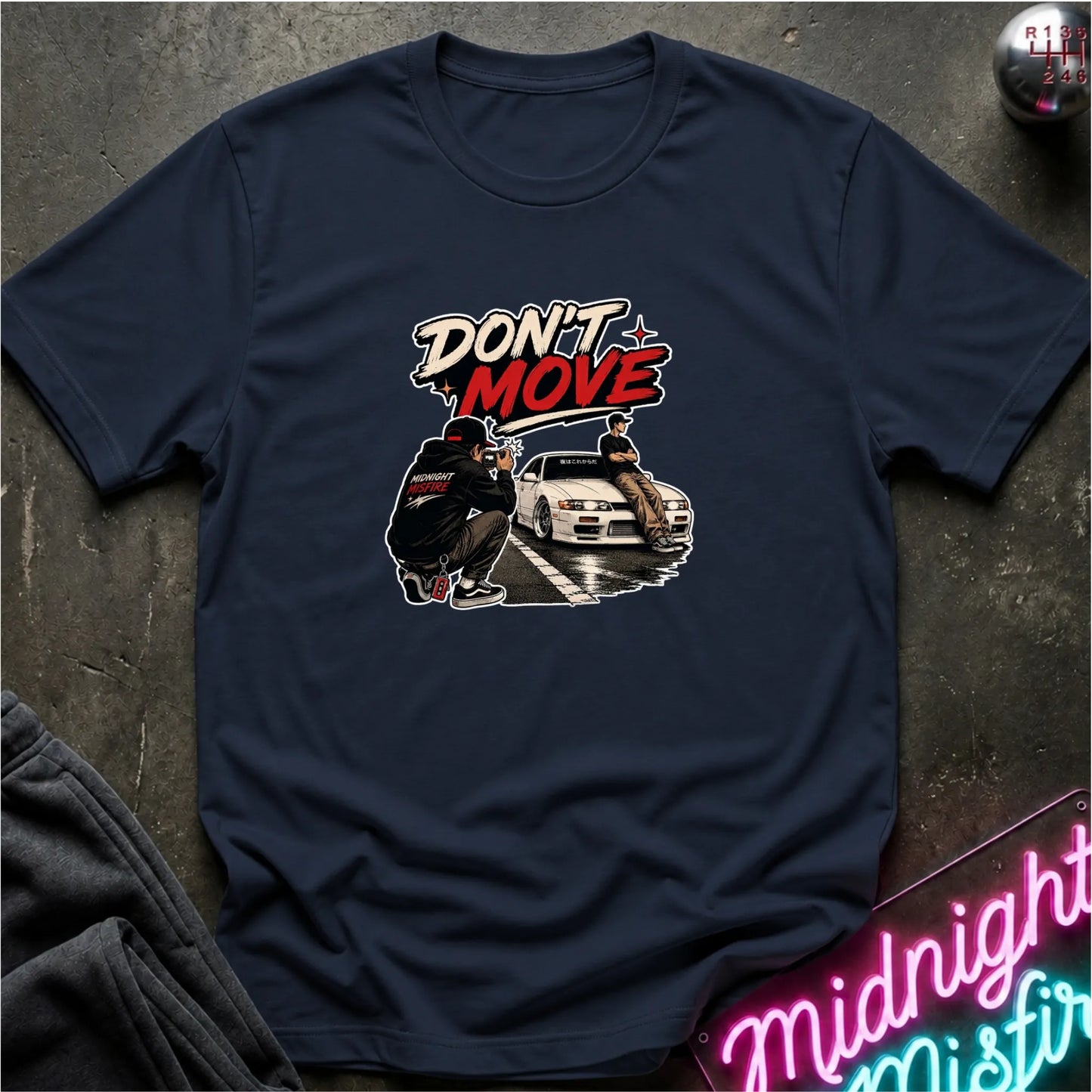 A navy blue graphic t-shirt laid flat on a dark textured surface. The graphic features the text 'DON'T MOVE' in stylized white and red lettering above an illustration of a white sports car and two figures.