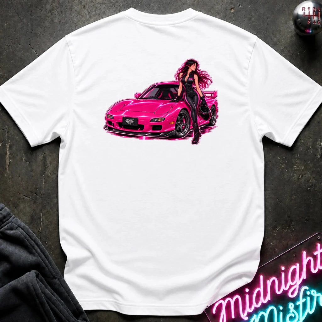 A white short-sleeve t-shirt laid flat on a dark textured surface, featuring a vibrant graphic print of a pink sports car with a woman in black standing beside it. The shirt is styled with a dark sweatshirt, a gear shift knob, and a neon sign in the background.