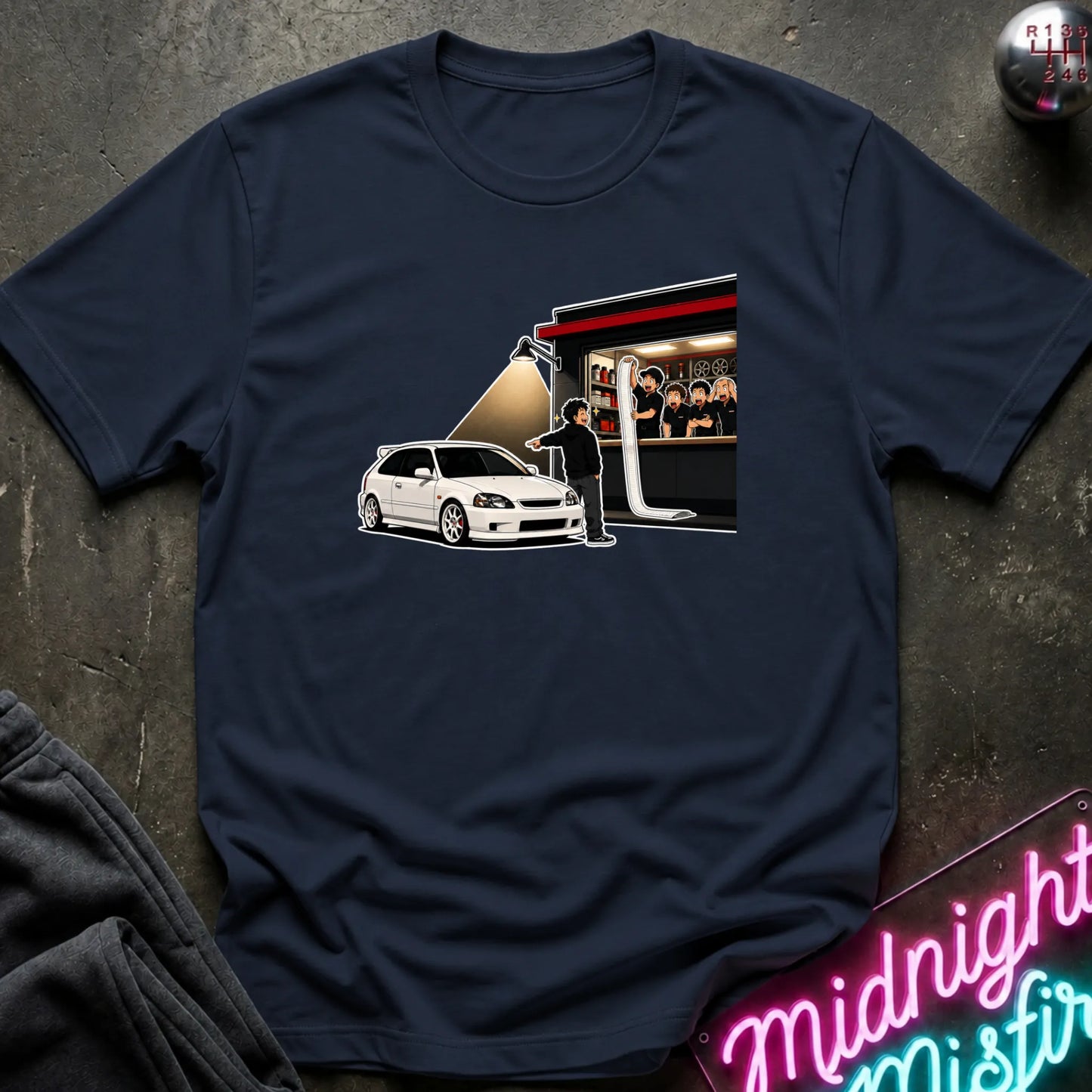 A navy blue short-sleeve t-shirt laid flat on a dark textured surface, featuring a graphic print of a white hatchback car in front of a stage with a spotlight and a group of people in the background.