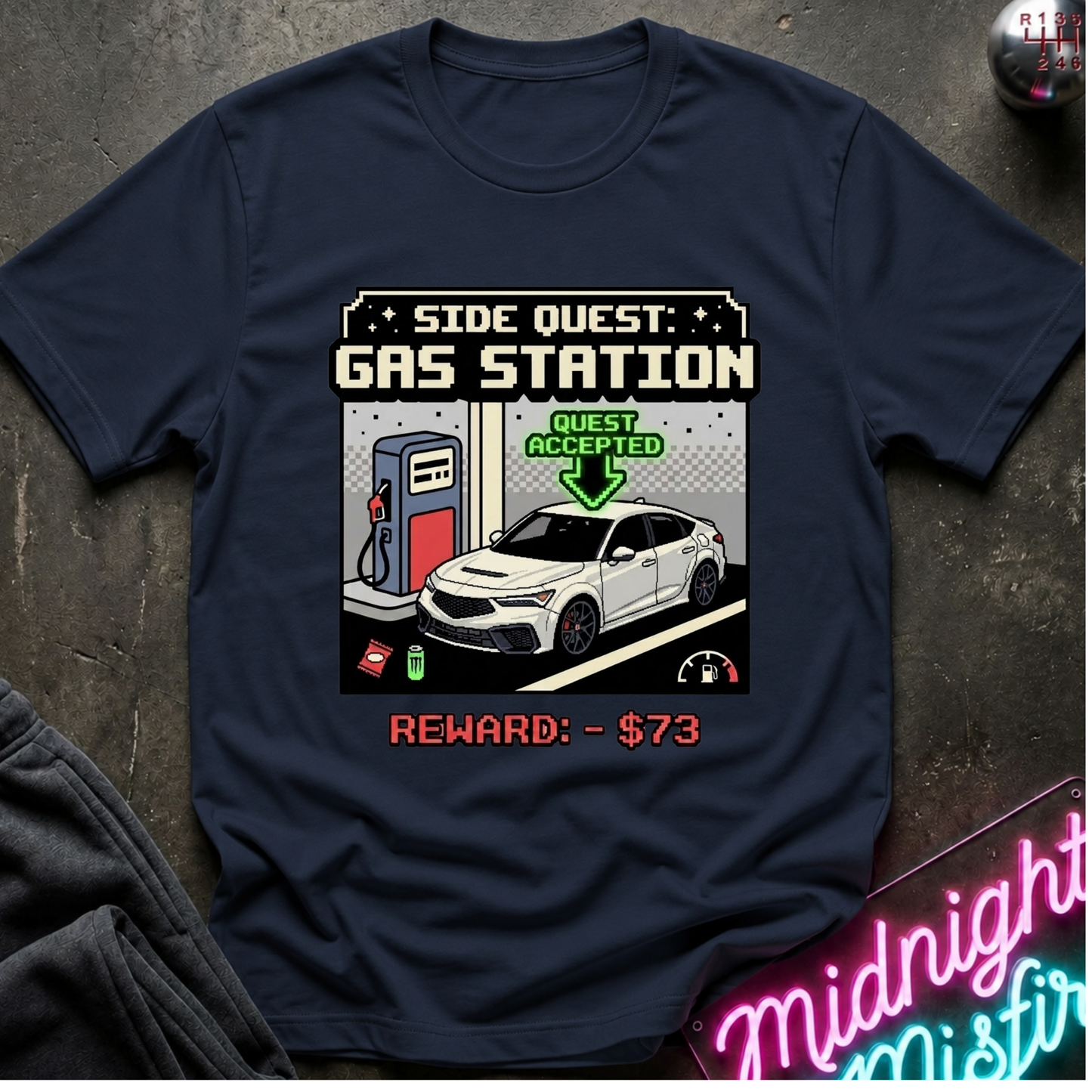 A navy blue graphic t-shirt laid flat on a dark textured surface. The shirt features a pixel-art style graphic of a white car at a gas pump with the text 'SIDE QUEST: GAS STATION', 'GUEST ACCEPTED', and 'REWARD: - $73'.