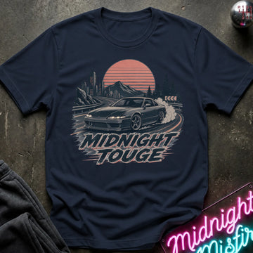 A navy blue graphic t-shirt laid flat on a dark textured surface. The shirt features a retro-style print of a black sports car drifting in front of a sunset over mountains, with the text 'MIDNIGHT TOUGHE' below.